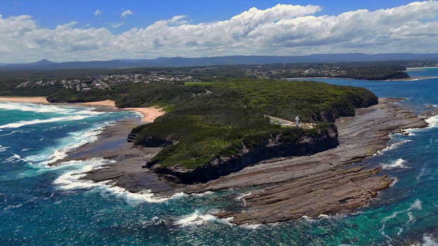 Sweeping establishing aerial of surf waves pounding dark rocks beneath Warden Head Lighthouse on the coastline, vibrant bright sunny day
