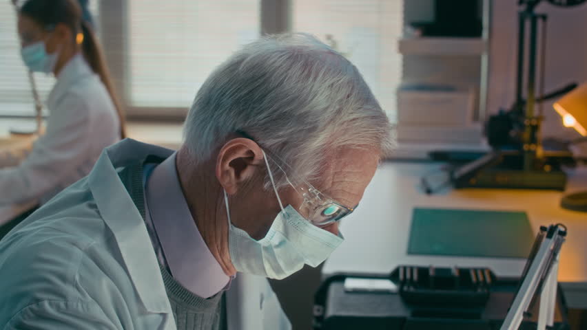 Arc slowmo shot of grey-haired male scientist in protective goggles and disposable face mask examining blue liquid substance in petri dish through magnifying glass, focused on research in laboratory