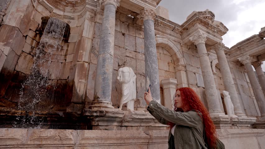 A female traveler filming the surrounding monumental stone sculptures and Roman architectural details in Sagalassos Ancient City, Burdur, Turkey. Cultural heritage, archaeology, historic architecture, and travel documentation concept.