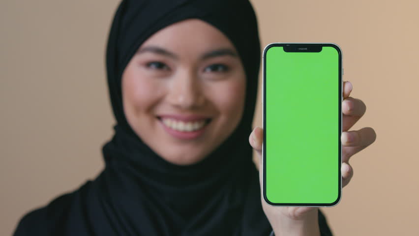 Smiling Asian Muslim woman in black hijab holding smartphone green screen looking at camera promo female showing mobile phone Islamic girl advertisement device online marketing technology app mockup