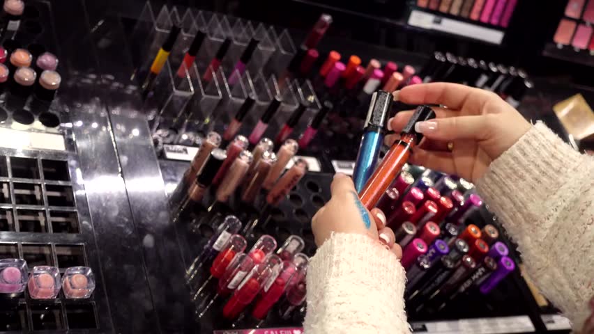 Woman shops for cosmetics in a mall, browsing lipstick and eyeshadow on shelves, highlighting beauty products and retail experience.