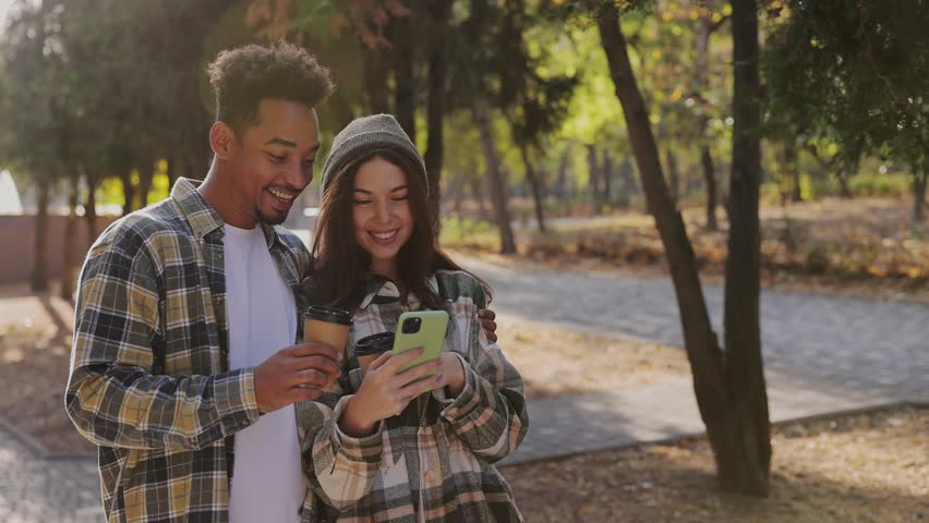 Young couple two friends man woman wear casual clothes together hold takeaway cup coffee use mobile cell phone walk rest relax in fall city park go down alley outdoors on nature. Urban leisure concept