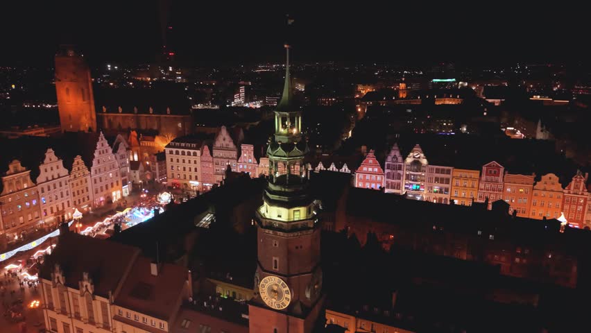 Aerial evening drone orbit around the tower of Wroclaw Town Hall, with the Christmas market visible below in the Main Market Square, showing festive stalls, holiday lights, and the historic Gothic architecture in the city center of Wroclaw, Poland.