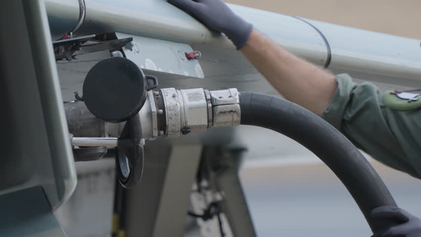 Military technician holding service hose connected to fighter jet to supply fuel or hydraulic fluid during controlled ground servicing procedure