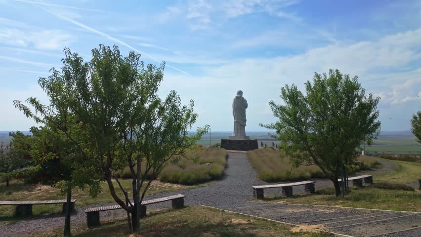 Sideways tracking reveals the Áldó Krisztus statue in Tarcal, Hungary, surrounded by lush greenery, open fields, and a peaceful park with pathways, benches, and a serene European landscape.