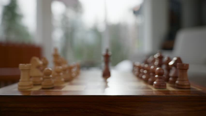 Zoom in as a hand makes a move with a piece on a chessboard while playing a game. The chess pieces are made of wood. The setting is a sunny room with a soft focus in the background.