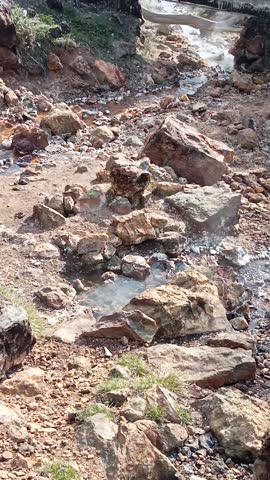 Steam rises from a natural hot spring with green hills and a clear sky in the background.