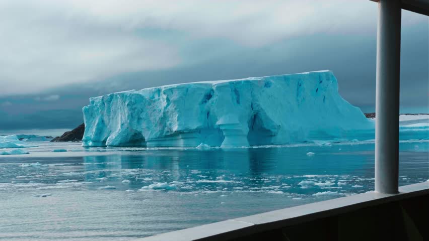 Small yacht sails between icebergs in the rays of sunset. Ship passes near huge ice cave in iceberg in Ilulissat, Greenland. Traveling on a yacht in the Arctic Circle, aerial shot