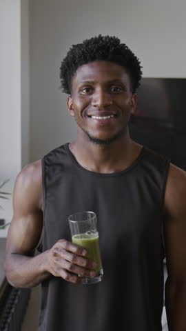 Vertical portrait of cheerful African American athletic guy posing for camera with glass of freshly blended smoothie