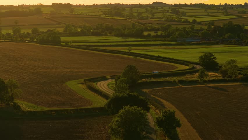 White car appearing on countryside curved road during glowing sunset. Car traveling countryside, road trip vacation, exploring sunset. Light vehicle advancing along rural winding roadway at golden