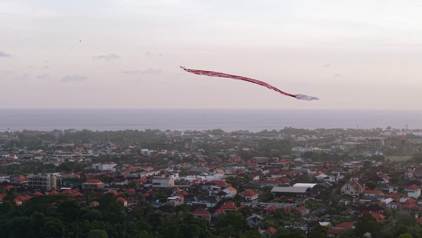 4K drone footage circling a large traditional Balinese kite soaring in the sky above Denpasar. The long tail flows through the air, celebrating a cultural festival rooted in local spiritual traditions.