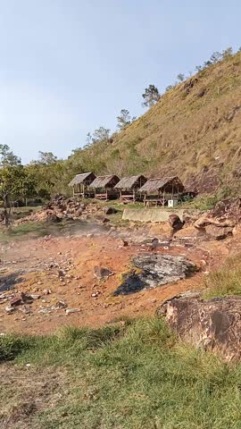 Steam rises from a natural hot spring with green hills and a clear sky in the background.