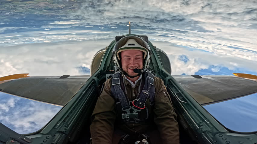 Professional male pilot in helmet and flight suit sitting in jet cockpit, military aviator flying plane high in blue sky above clouds