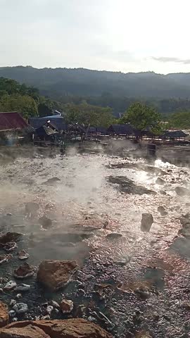 Steam rises from a natural hot spring with green hills and a clear sky in the background.