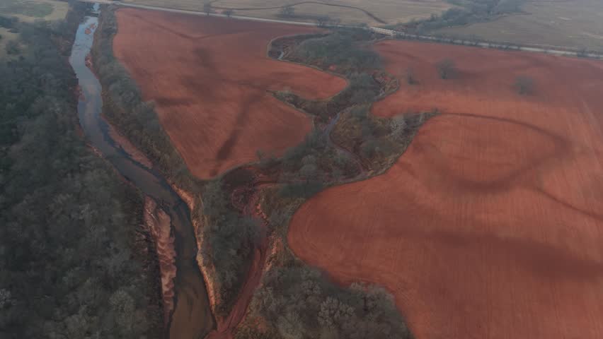 Aerial view of red soil farmland with plowed agricultural fields divided by a winding creek and tree-lined waterway in a rural landscape.