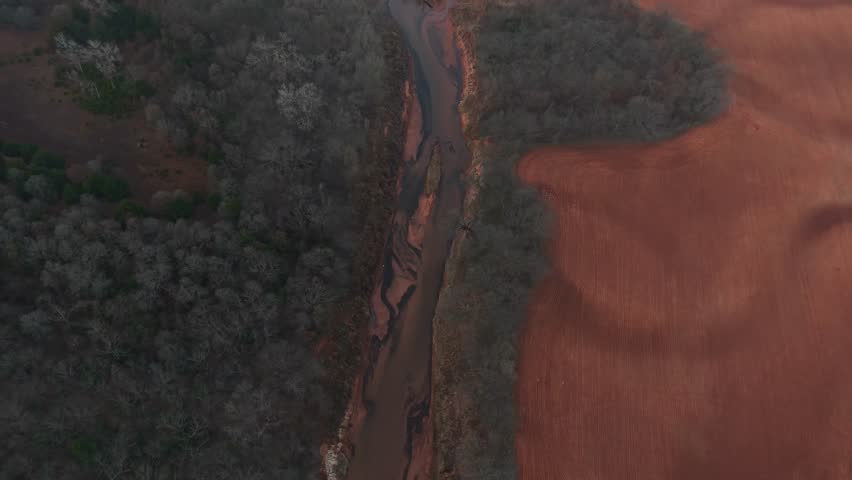 Aerial view of a narrow river winding between dense woodland and open farmland, showing contrasting natural forest and cultivated fields from above in a rural landscape.