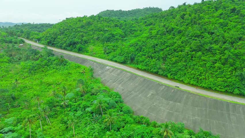 Aerial concrete highway winding through dense green tropical jungle and hills, with motorcycles traveling along the road in Siargao, Philippines.