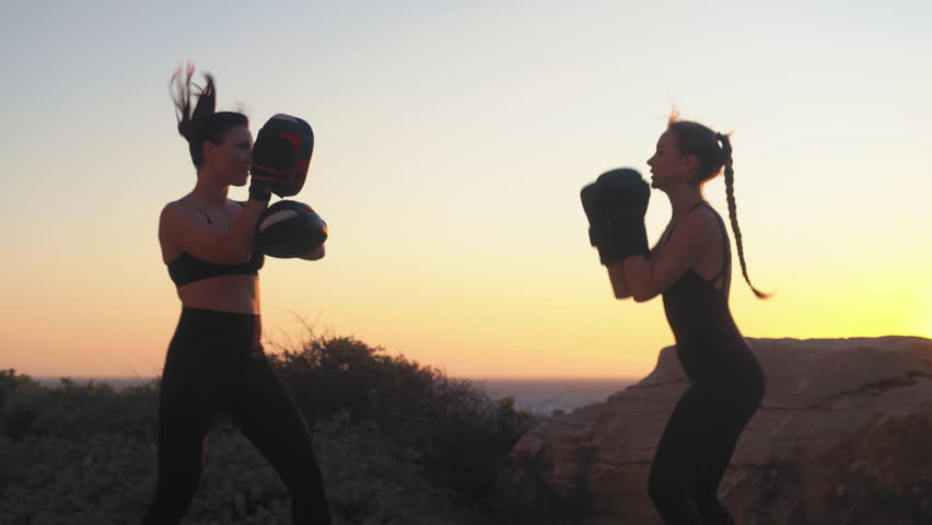 Sporty female fighter practicing kickboxing on hill overlooking seascape at sunset, with female coach guiding during focused training session in scenic beach setting