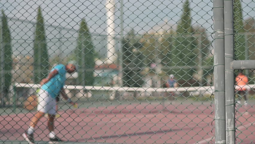 A group of tennis players captured out of focus while practicing on an outdoor tennis court. The foreground shows a chain-link fence in sharp detail, creating a cinematic and abstract sports atmosphere. Ideal for themes of training, motion, anonymity, sports lifestyle, competition, and outdoor fitness.