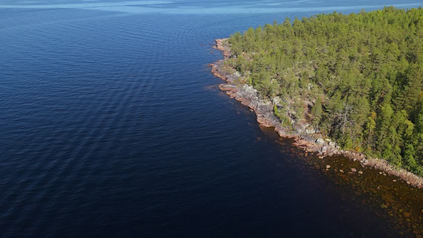 A rocky forested shoreline extends into a deep blue lake, with calm rippling water filling most of the frame and dense green trees covering the sunlit land on the right side. A bird-eye view of a