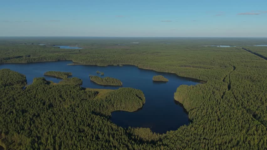 Aerial view of a dark blue lake with several forested islands, surrounded by dense green woodland stretching to the horizon under a clear blue sky.
