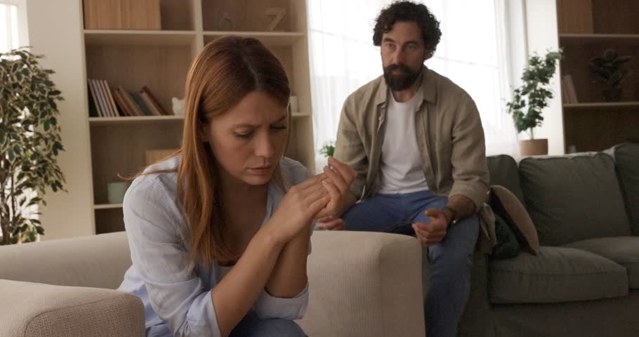 A distressed woman sits on the sofa, ignoring her frustrated husband during a serious argument, highlighting marital problems and relationship challenges, reflecting a deep emotional conflict.