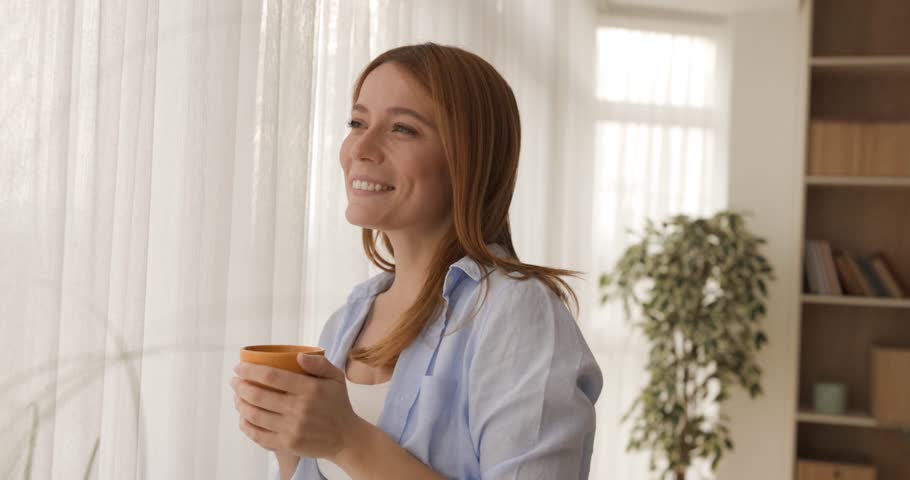 A cheerful young woman savors her morning coffee, looking out the window with a warm smile. This tranquil moment of relaxation and peace captures a cozy, happy start to the day at home.