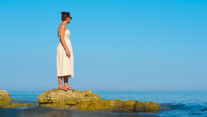 Woman in white dress standing on a rock in the sea and enjoying the view. Young woman in a white summer dress and sunglasses standing on a rock in the middle of the sea, enjoying the warm sunlight