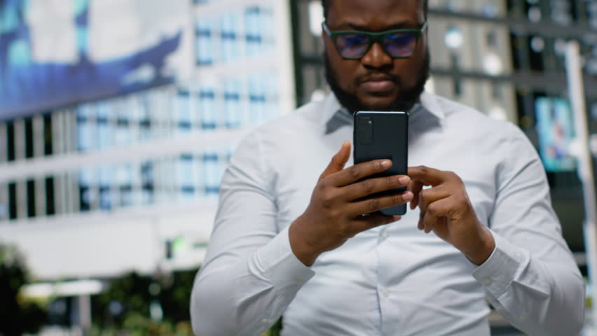 Black man looking for directions on smartphone map app during a stroll, walking through urban street with office buildings. Vibrant city life within a metropolitan city center area. Camera B.