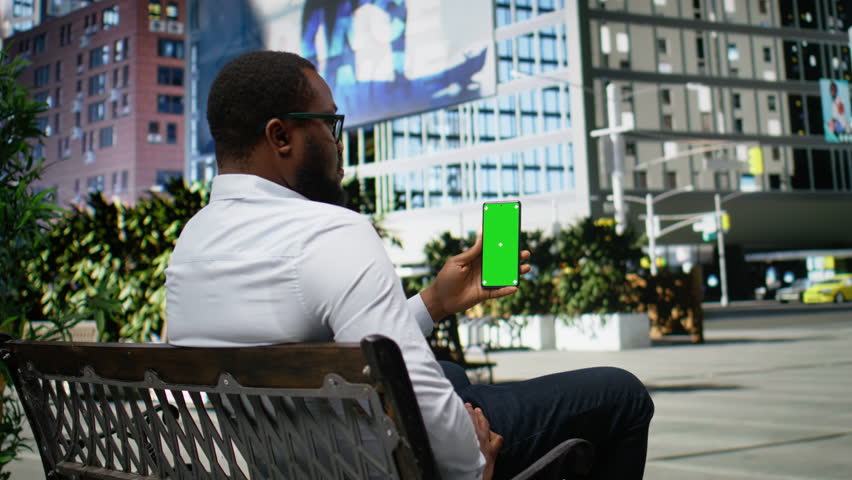 Vertical green screen with male sitting on a bench in a trendy city area, navigating web content with motion as his street style and attitude reflect energy of metropolitan urbanism. Camera A.