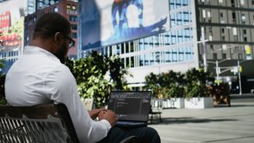 Confident adult reviews binary code script for his programming remote work, sitting on a bench in the city center and working on laptop. Scrolling and typing near lively downtown scenery. Camera A. - Powered by Shutterstock - Get 15% off with code: PIKWIZARD15