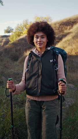 Happy African American woman with Nordic walking poles stands in ravine among hills at sunset. Sportive black female tourist rests on hike at hilly countryside