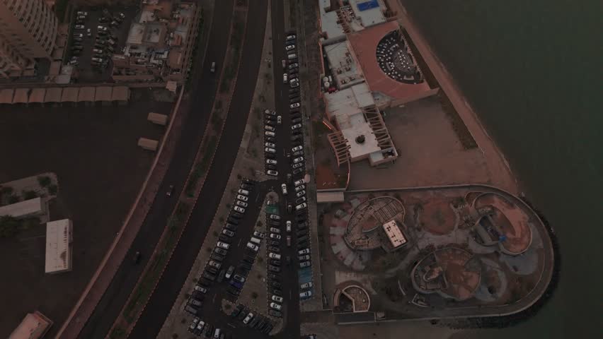 Top-down drone of a coastal city block: seafront venue with curved terrace, packed parking rows, divided roadway, shoreline and dark water; blue hour evening light, no people—great for travel or urban