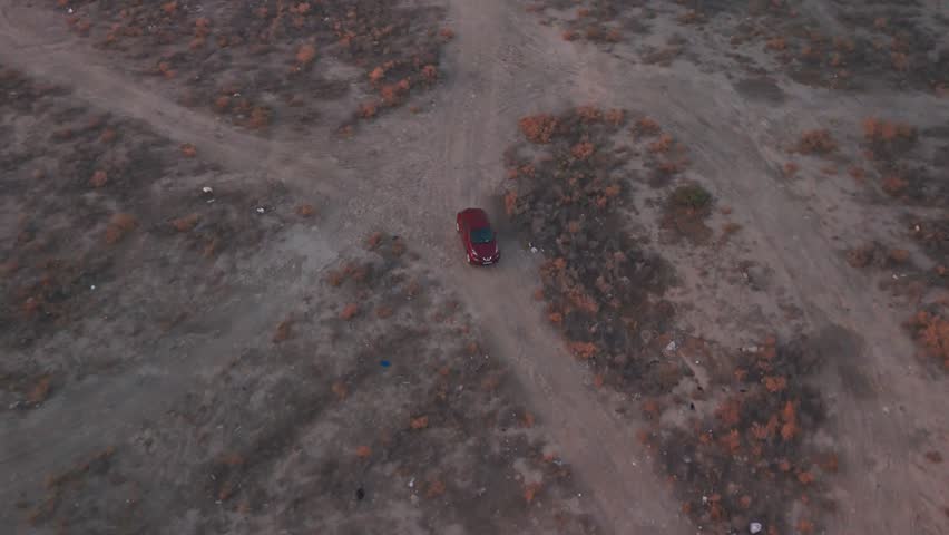 Top-down drone of a red SUV driving along a desert dirt road through scrub; wide barren terrain, evening light, tiny distant figures—ideal for travel, off-road, and adventure B-roll.