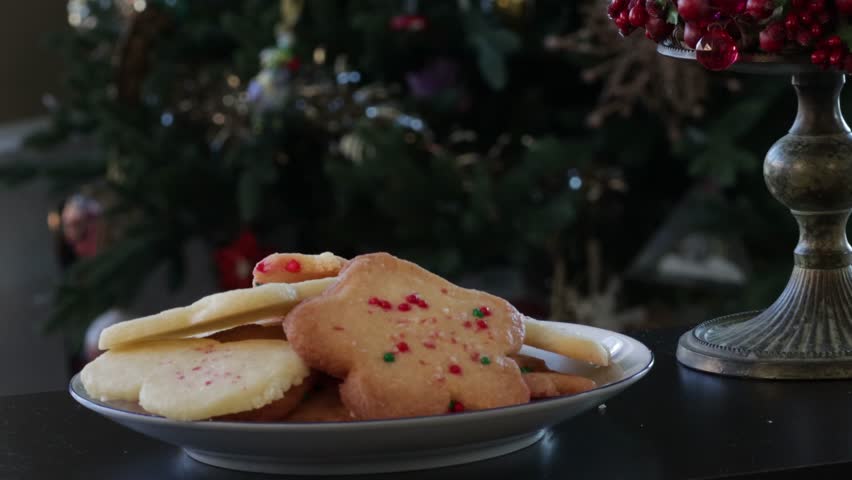A close up of a hand reaching down to a plate of decorated cookies and sneakily taking one. A Christmas tree and other holiday decorations are in the background.