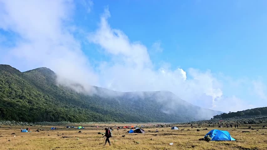 People passing by in mountain campsite in Surya Kencana, Gede-Pangrango Mountain, West Java, Indonesia. Camping tents in a savanna with mountainous background and clear sunny sky.