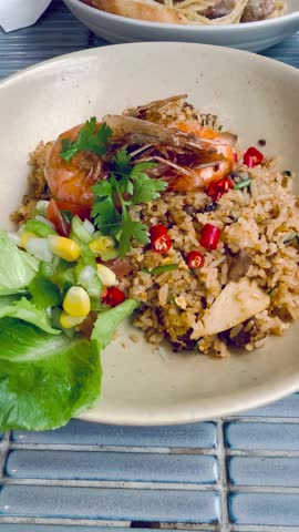 A close-up view of a plate of tom yum fried rice (Nasi Goreng Tom Yum) with prawns, chili slices, cilantro leaves, and salad.