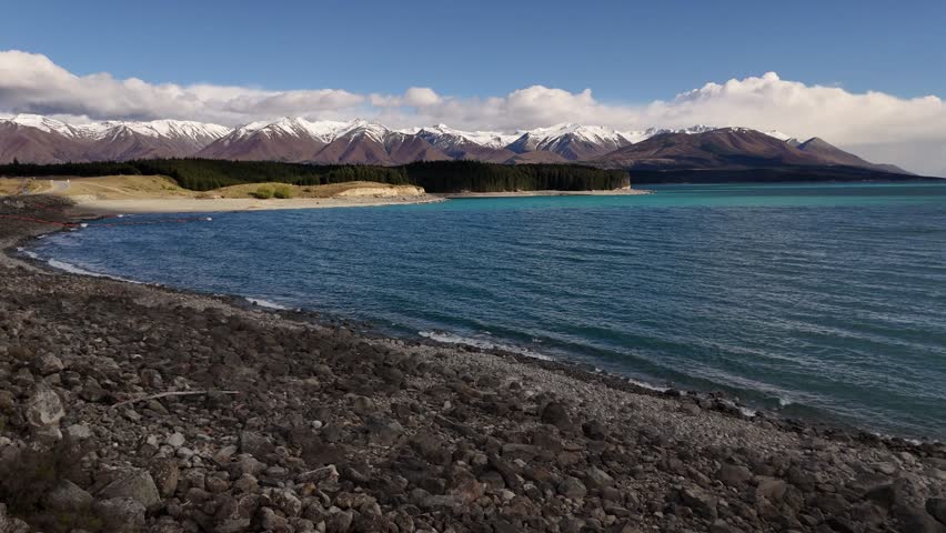 Rocky shoreline at Lake Pukaki with turquoise water and snow-capped Southern Alps in the background. Clear day in New Zealand’s South Island. Aerial wide shot.