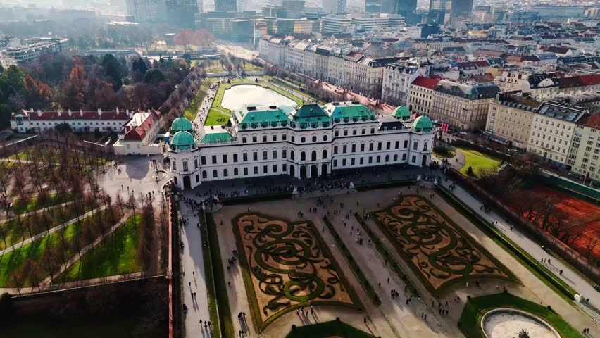 Aerial drone shot quickly panning left over Belvedere Palace in Vienna, capturing the baroque palace and gardens in the foreground with the winter Vienna cityscape visible in the background under sun