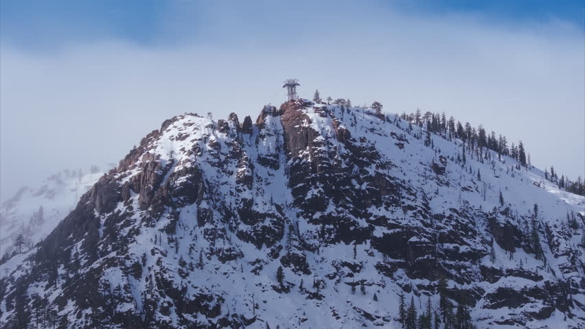 Aerial view of snowfall covering rocky mountain summit at Palisades Tahoe, California, USA. Snowy cliffs, alpine terrain and evergreen trees create dramatic winter landscape in Sierra Nevada mountains
