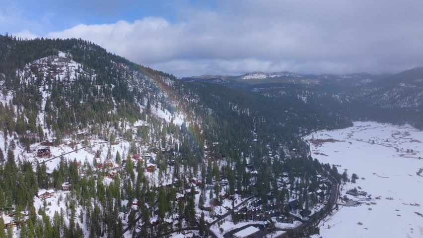 Aerial view of light snowfall over forested mountain valley at Palisades Tahoe, California, USA. Snow covered cabins, pine trees and hills appear beneath soft rainbow, forming a calm winter landscape.