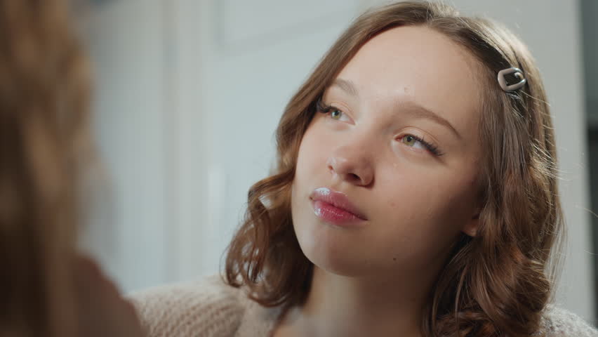 CloseUp Of Young Woman Rehearsing Expressions While Makeup Is Applied, Hairclip And Sweater Present, Quiet Studio Rehearsal Vibe With Focused Gaze And Subtle Cosmetic Touches For Portfolio Readiness