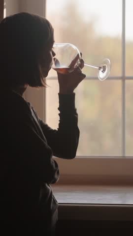 Elegant young woman drinking wine from a beautiful glass, standing in backlight on a modern kitchen. Female silhouette by the window creates a calm, cinematic evening lifestyle mood.
