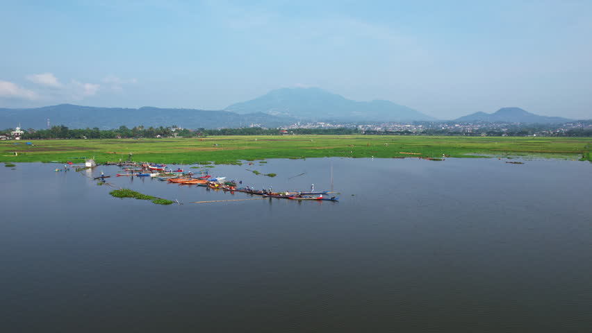 Aerial drone footage of a local fishermen waiting to board their wooden traditional boats in a water reservoir in Indonesia, with paddy and mountain background