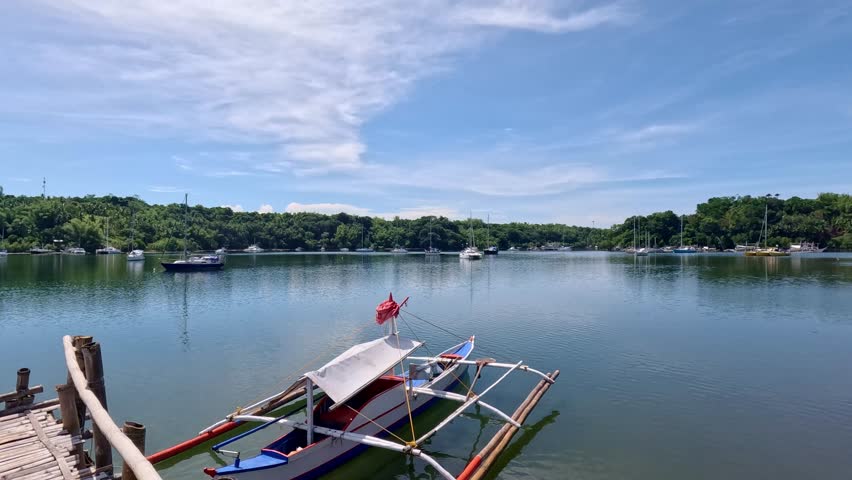 Pan left showing an outrigger boat moored beside a bamboo boardwalk and anchored sailboats in the distance at Tambobo Bay. Siaton, Negros Oriental, Philippines