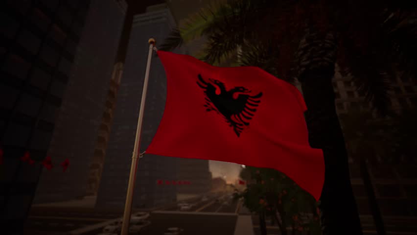 The Albanian national flag waves gently at sunset along a modern city street, illuminated by warm golden light that reflects across surrounding buildings and urban surfaces. The contrast between the deep red of the flag and the warm tones of the sky creates a calm and visually balanced atmosphere. The black double-headed eagle remains clearly visible, reinforcing the flag’s symbolic presence.

Urban elements such as streets, buildings, and light traffic appear in the background without overpowering the composition, keeping the flag as the central focus. The absence of recognizable landmarks allows the image to function as a general representation of Albania rather than a specific location. Subtle motion in the flag adds realism while preserving a composed and professional tone.

This visual is suitable for editorial use related to international news, European affairs, cultural storytelling, or travel features. Its warm lighting and neutral composition provide flexibility for a wide range of informational and media applications.
