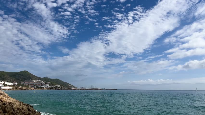 View of Passeig de la Ribera in Sitges with turquoise Mediterranean, sandy beach, white coastal buildings and sailboats under a blue summer sky.