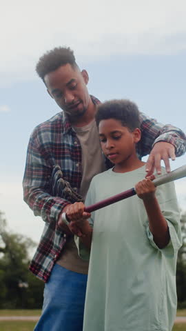 Confident man standing behind black boy and teaching holding baseball bat in right places and comfortable position. Sports trainer practicing athletic skills and techniques with interested offspring.
