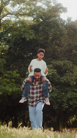 Full length of playful parent carrying little offspring on shoulders and kicking soccer ball up to child above. Loving family having fun at yard with trees and entertaining with equipment for game.