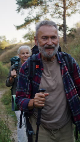 Happy old man with wife walks using canes between low green hills. Cheerful mature man and woman travel practicing Nordic walking at spring countryside